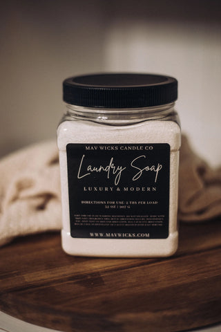 A jar of "Laundry is SO much fun when it smells this great!!!" from Mav Wicks Candle Co. features a black label that reads "Luxury & Modern." The directions suggest using 1-2 tablespoons per load for deep cleaning. The jar is displayed on a wooden surface with a folded cloth in the background.