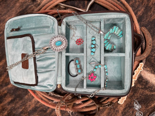 The Carson Cowhide and Tooled Leather Travel Jewelry Case by American Darling, crafted in teal genuine leather and featuring a YKK zipper, is filled with various pieces of turquoise jewelry such as necklaces, bracelets, earrings, and rings. Each piece exhibits intricate silver detailing. The case is displayed on a rustic wooden surface with coiled rope beside it.