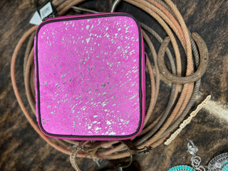 The Poesy Cowhide and Leather Travel Jewelry Case by American Darling, featuring a brightly colored square pink design crafted from Genuine Full Grain Leather with a silver speckled pattern, is elegantly placed atop coiled ropes and a horseshoe. Partially visible to the right is a silver accessory adorned with turquoise accents. The background showcases a textured, rustic surface.