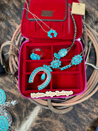 An open red Poesy Cowhide and Leather Travel Jewelry Case by American Darling showcases turquoise and silver jewelry, featuring necklaces, earrings, a pendant, and a ring. The pieces highlight floral and horseshoe designs, with coiled rope elements and Genuine Full Grain Leather accessories in the background.