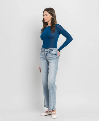 A woman stands against a plain white background, wearing a blue long-sleeve top and the Riggs Mid Rise Tummy Control Mid Rise Wide Leg jeans from Western Edge Boutique. With her brown hair flowing and her gaze directed to the side, she completes her casual, relaxed look with white shoes.