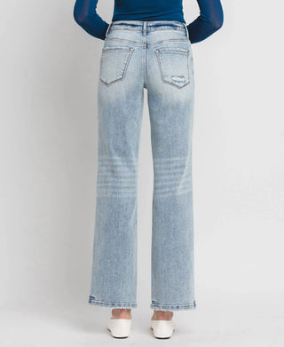 A person is wearing the Riggs Mid Rise Tummy Control Wide Leg jeans from Western Edge Boutique, featuring a light blue wash and subtle distressing on one back pocket. They have paired them with a dark blue long-sleeve top and white shoes, standing against a plain white background.