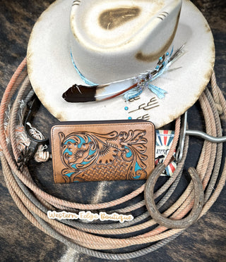 A white cowboy hat adorned with a feather and turquoise bead embellishment is placed atop the Etta Tooled Leather Wallet, an exquisite piece by American Darling. This decorative wallet is made from harness skirting leather and showcases intricate hand-carved floral and basketweave patterns. Beneath them lie intertwined ropes, a horseshoe, and a branded tag that reads "Western Edge Boutique." SIZE: H 4.5" X W 8".