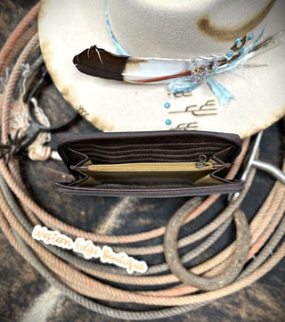 An Etta Tooled Leather Wallet by American Darling, crafted from harness skirting leather in brown, is open and displayed on a rustic background. It is surrounded by coiled ropes, a horseshoe, and a cream-colored cowboy hat adorned with feathers and turquoise beads. A "Western Edge Boutique" tag is partially visible at the bottom. SIZE: H 4.5" X W 8".