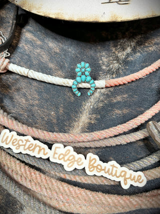 Felicity Squash Adjustable Ring, featuring an elegant design with multiple small turquoise stones, is displayed on coiled ropes. Below the ring, there's a wooden plaque engraved with "Western Edge Boutique." The rustic background surface enhances the charm of the burnished silver accents and vibrant turquoise color.