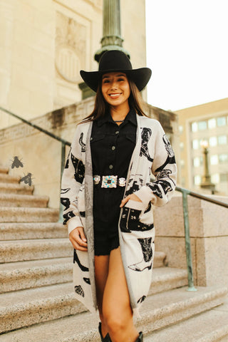 A woman in a black hat and outfit stands on stone steps, her "Boots a Million Cardigan" by 2FLY adding flair with its graphic design and decorative belt buckle. Urban buildings loom behind, completing this cozy scene.