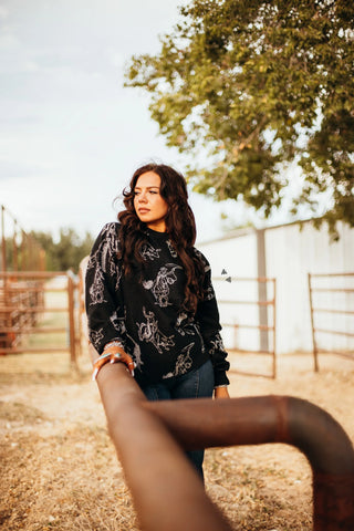 A person with long hair in a Sparkle Buck Black Silver Metallic Sweater by 2FLY leans on a rusty fence. Trees and a white building under a cloudy sky add western flair to the scene.