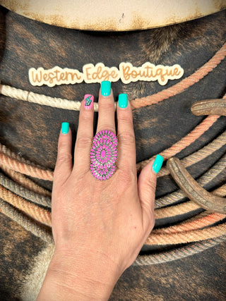 A hand with turquoise-colored nails and a decorative accent nail is shown wearing the Sharna Western Concho Stretch Ring from Western Edge Boutique. This large, vibrant pink ring boasts exquisite burnished silver details, giving it a unique flair. Coiled ropes and the Western Edge Boutique sign are visible in the background, enhancing the rustic, Western theme.