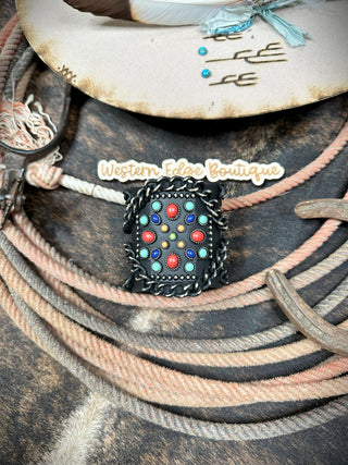 A vintage-inspired western bracelet with turquoise and red colored stone inlays is displayed on coiled rope and a rustic cowboy hat. Crafted from burnished silver, the Jaedan Western Cross Cuff Bracelet shines beautifully. The background features a "Western Edge Boutique" sticker.