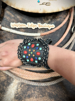 A wrist adorned with the Jaedan Western Cross Cuff Bracelet, a wide black leather accessory featuring an intricate design of colorful stones including turquoise, red, and yellow. The bracelet boasts braided edges and hints of burnished silver. In the background, there's a sign that reads "Western Edge Boutique" with ropes as decor.