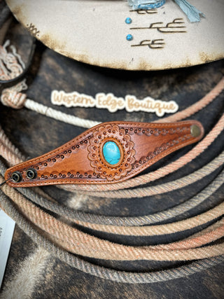 A close-up of a handcrafted Sienna Leather Snap Bracelet made from cowhide leather, featuring an intricate pattern and a turquoise stone in the center. The background showcases coiled ropes and an "American Darling" label. Small turquoise beads and Western-themed designs are also visible, highlighting the charm of this Western style leather bracelet.