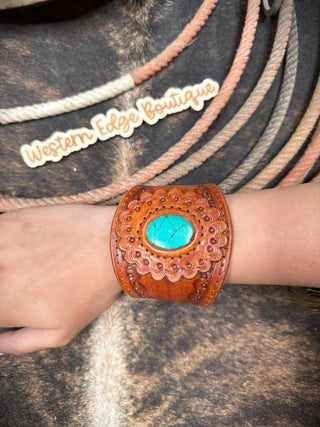 A wrist showcasing the Sienna Leather Snap Bracelet by American Darling, a wide, brown handmade leather bracelet with an intricate cutout design and a central turquoise stone. The background includes coiled ropes and a "Western Edge Boutique" sign in a rustic style.