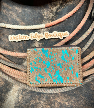 A small Piper Hair-on Hide Leather Zipper Pouch with turquoise speckles lies on top of coiled ropes. Above the pouch is a stylized "American Darling" sign. The background features a textured surface that resembles hair-on cowhide genuine leather, highlighting the rustic charm and durability of our accessories.