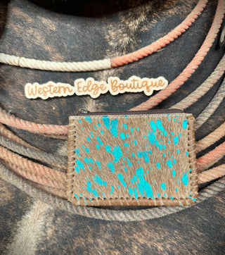 A small Piper Hair-on Hide Leather Zipper Pouch from American Darling, crafted from genuine hair-on cowhide leather with brown and turquoise speckles, is placed on a background of coiled ropes. Above the pouch is a wooden sign with the engraved text "Western Edge Boutique." The pouch features durable zipper closures to secure your coins and credit cards.