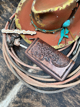 The Sedona Tooled Leather Wallet by American Darling, featuring intricate embossing, is displayed alongside a worn cowboy hat adorned with turquoise embellishments, a coiled lasso, and a Western Edge Boutique tag. The items rest on a brown and white animal hide backdrop, embodying an authentic western look.