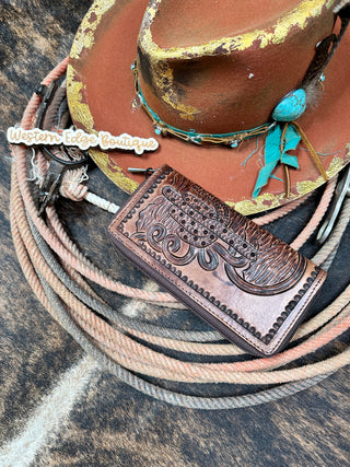 A perfectly crafted Sedona Tooled Leather Wallet by American Darling in brown Full Grain Leather, featuring intricate tooled designs, lies next to a rustic cowboy hat adorned with a turquoise arrowhead decoration and a rope lariat. Completing the Western look on a themed backdrop, the image also includes a "Western Edge Boutique" tag.