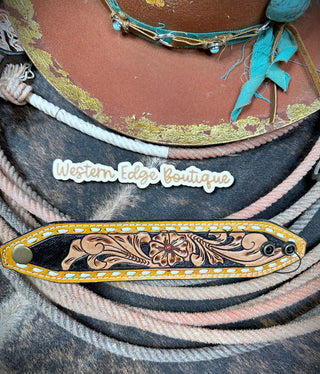 A close-up of the Lark Tooled Buckstitch Snap Bracelet from American Darling, showcasing its decorative, hand-tooled leather strap with intricate floral carvings, set against a patterned background. Above the strap, there's a logo that reads "Western Edge Boutique." Various cords and threads are also visible, adding texture and color to the Western style image.