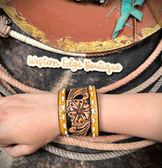 A close-up of a wrist adorned with the American Darling's Lark Tooled Buckstitch Snap Bracelet, featuring intricate floral carvings and yellow accents. The background displays intertwined ropes and feathers against a "Western Edge Boutique" sign, enhancing the Western style ambiance.