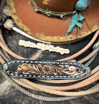 A close-up showcases the intricate floral tooling on the Paige Buckstitch Leather Snap Bracelet by American Darling. In the background, a brown hat adorned with turquoise accents and beaded embellishments rests atop tangled ropes. A sign for "Western Edge Boutique" is partially visible in the distance.