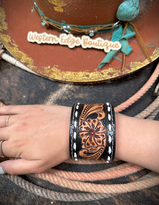 A hand wearing the Paige Buckstitch Leather Snap Bracelet, featuring a floral pattern in brown and black with a red bead, is shown. In the background, a brown hat adorned with turquoise and leather accents from Western Edge Boutique rests on a textured surface next to coiled rope.