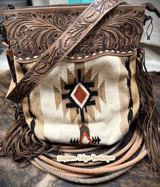 The Mesa Saddle Blanket and Tooled Leather Handbag by American Darling showcases intricate floral leatherwork on the top and strap, complementing its woven fabric body adorned with a geometric Southwestern pattern. Made from genuine full grain leather and hand-woven woolen saddle blanket, this stylish handbag also features fringe details and proudly bears the "Western Edge Boutique" label.