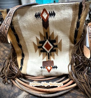 The Mesa Saddle Blanket and Tooled Leather Handbag by American Darling is displayed in a crossbody style featuring a Southwestern design in beige, black, and rust patterns. It showcases intricate fringe details on the sides and boasts a tooled leather strap made from genuine full grain leather. Near the bottom is a small leather tag that reads "Western Edge Boutique." The background is set against a rustic wooden surface.