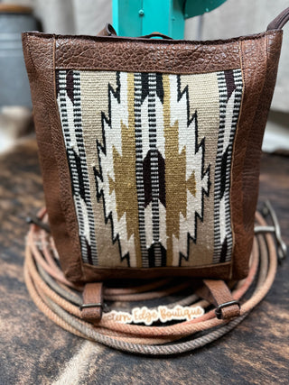 The Misty Saddle Blanket and Leather Concealed Carry Handbag by American Darling is a stylish brown leather handbag crafted from Full Grain Leather, featuring an eye-catching Native American-inspired geometric pattern in black, white, taupe, and beige at its center. Photographed against a rustic wooden background with coiled ropes at its base, it boasts durable zipper closures. A tag reads "Western Edge Boutique.