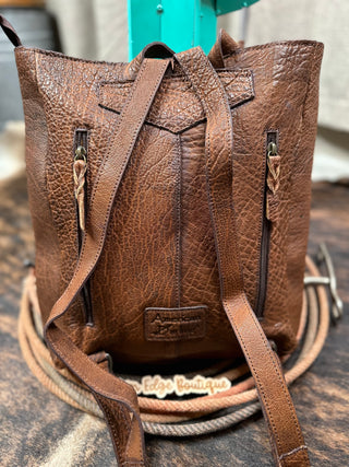 A rugged brown leather Misty Saddle Blanket and Leather Concealed Carry Handbag made from Full Grain Leather with a textured finish is displayed on a coiled rope. The handbag features two vertical front zippers and durable zipper closures on the adjustable straps. A tag with the label "American Darling" is attached at the bottom.