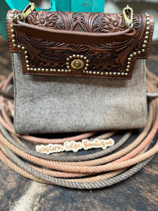 Presenting the Roamer Cowhide and Tooled Leather Concealed Carry Handbag by American Darling: This stunning piece features a tooled leather flap embellished with brass studs and a secure latch closure. The bottom half is crafted from light tan genuine leather, giving it an elegant yet rugged appeal. Displayed on coiled ropes, it perfectly complements the Western Edge Boutique backdrop.