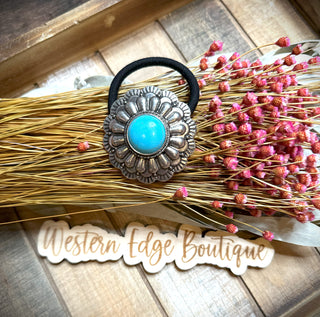 A decorative Heidi Concho Hair Band with a turquoise stone set in an ornate metallic flower design is placed atop a bundle of dried wheat and pink flowers. Below, "Western Edge Boutique" is written in a cursive font on a wooden surface.