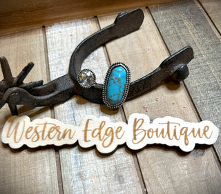 A stunning Patty Turquoise Cuff Ring, featuring a large turquoise stone and clear colored accents, is displayed elegantly on an old metal horseshoe against a rustic wooden backdrop. Prominently in the foreground, a wooden sign bears the name "Western Edge Boutique."
