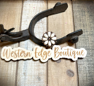 A rustic, weathered wood background features a metal horseshoe wall hook. Below the hook is a wooden sign with the text "Western Edge Boutique" in elegant script. Atop the hook sits the Kady Flower Cuff Ring adorned with white stones and subtle copper accents.