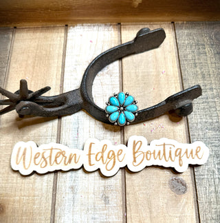 A Florence Flower Cuff Ring with burnished silver accents is displayed on a rustic wooden surface next to a wrought iron horseshoe. Below the ring and horseshoe, there is a sign with the inscription "Western Edge Boutique" in a stylish cursive font.