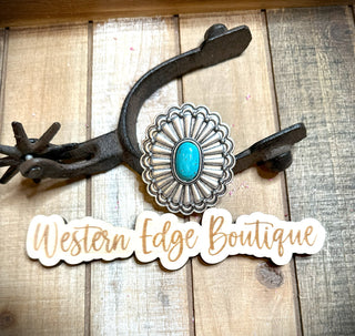 A Hallow Turquoise Concho Adjustable Ring is displayed on an antique silver plate next to a rustic spur. Below, a sign reads "Western Edge Boutique" in stylish script, all arranged on a wooden surface.