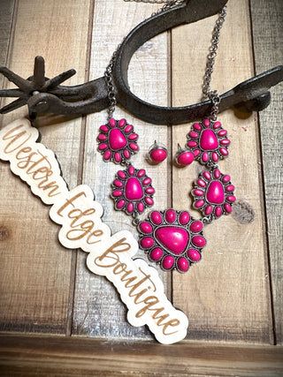 A close-up of the Patrice Concho Necklace Set, featuring striking fuchsia stones set in burnished silver frames, displayed against a rustic wooden background. A metal horseshoe is in the background, and a wooden sign reads "Western Edge Boutique".