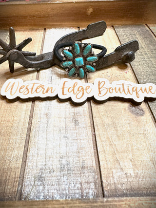 A Sarah Concho Hair Tie, featuring turquoise gemstone accents and burnished silver details, is elegantly displayed on a wooden surface. Below it, a rustic sign with "Western Edge Boutique" written in stylish script font completes the Western-themed aesthetic.