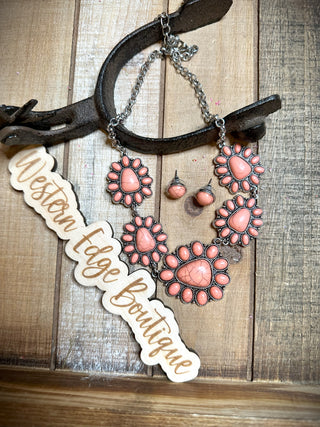 A jewelry set featuring the Priscilla Concho Necklace, matching earrings, and bracelet adorned with pink stones arranged in floral patterns is displayed on a rustic wooden wall with an antler hook. A wooden sign with cursive text reads "Western Edge Boutique.