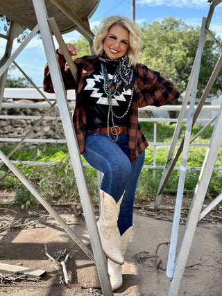 A person wearing a black patterned shirt, the Everyday Comfort Flannel from Western Edge Boutique, jeans, cowboy boots, and a bandana poses outdoors against a metal framework, with greenery and a water tower accenting the scene.