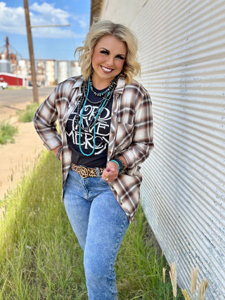 A smiling woman stands outdoors next to a corrugated metal building. She is wearing a Carmel Clay Flannel Top by TEXAS THREAD over a graphic tee, jeans, a belt with a large buckle, and multiple turquoise necklaces. The fall color combo complements the background of a road, grass, and distant industrial structures.