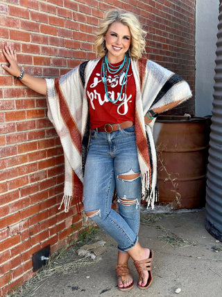 A woman stands against a brick wall, smiling. She wears a red shirt with white text, the Salem Plaid Kimono by TEXAS THREAD, ripped blue jeans, and brown sandals. She has layered necklaces and bracelets, and her hair is styled in waves. She leans on the wall with one hand.