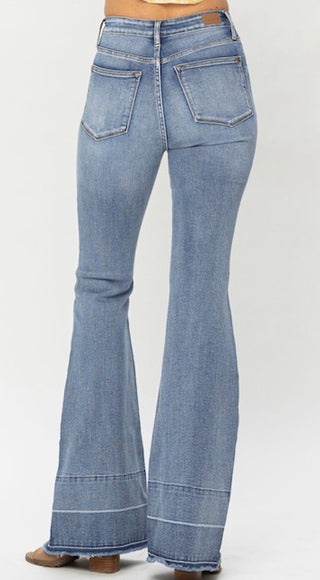 A person wearing light blue Townes High Waisted Tummy Control Judy Blue Flares from Western Edge Boutique is shown from behind. The jeans showcase two back pockets with stitching details and wide flared legs. The person is also sporting brown suede shoes.