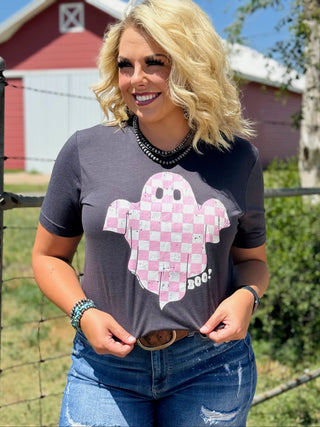 A woman with curly blonde hair stands outdoors, smiling. She is wearing the must-have Hey Boo Graphic Tee Halloween Graphic Tee from Western Edge Boutique, featuring an adorable pink checkered ghost with the word "BOO!" written on it. She pairs this outfit with distressed blue jeans, a belt, bracelets, and a beaded necklace. A barn is visible in the background.