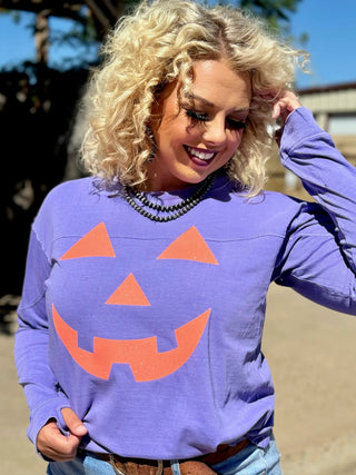 A person with curly blonde hair smiles while wearing a lavender Glitter Jack O'Lantern Long Sleeve Top from Western Edge Boutique. They are standing outside on a sunny day, with trees and a building visible in the background, fully embracing the Halloween festivities.