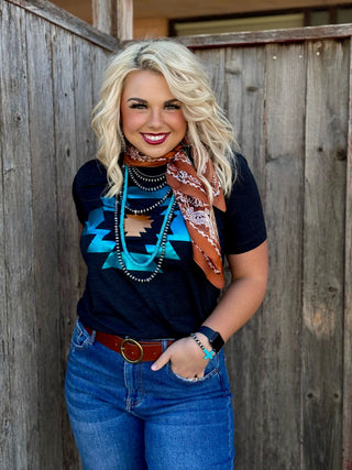 A woman with blonde hair is smiling and standing in front of a wooden wall. She is wearing the Durango Aztec in Turquoise and Copper Foil tee by Western Edge Boutique, blue jeans, a brown belt, multiple necklaces, a brown bandana around her neck, and a smartwatch on her left wrist.