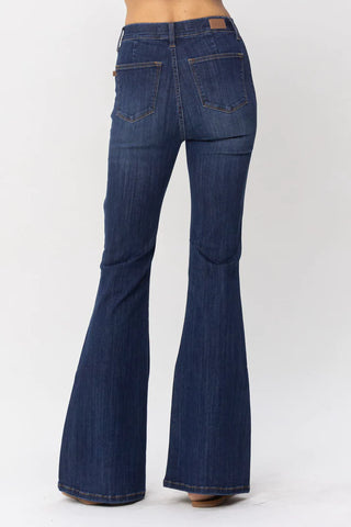 A person is modeling a pair of dark blue Roe Flare Jeggings from Judy Blue. The jeggings feature back pockets and are fitted through the hips and thighs, flaring out from the knees down. The person is standing with their back facing the camera against a plain white background.