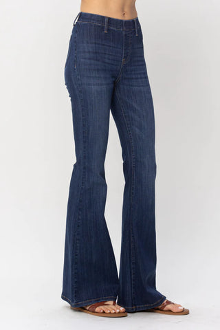 A person is wearing dark blue, high-waisted Roe Flare Jeggings by Judy Blue. They are standing sideways against a plain white background. The jeggings fit snugly around the hips and thighs and flare out from the knees. The person is wearing flat, open-toe sandals.