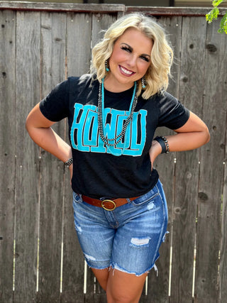 A smiling person stands in front of a wooden fence. They have short blonde hair and are wearing a Howdy Black Turquoise Tee from Western Edge Boutique, featuring bold turquoise letters spelling "Howdy." They are also wearing denim shorts, a brown belt, and matching turquoise jewelry.