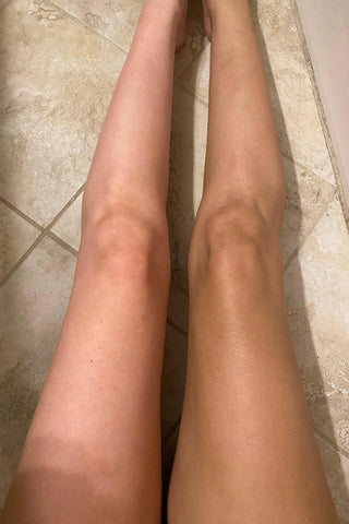 On a tiled floor, two legs are displayed side by side, with the left leg noticeably lighter and the right darker, illustrating the difference achieved using Western Edge Boutique's vegan-friendly Self Tanner Mousse in Medium Shade.