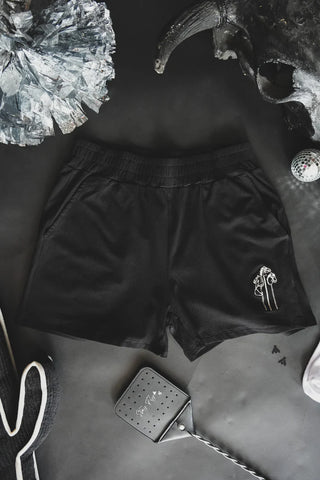 The LASSO THE COMFY SET by 2FLY lays on a flat, dark surface, featuring black lounge wear shorts with a small stick pony embroidery near the left pocket, surrounded by a glittery silver pom-pom, a shiny black and silver mask, a silver disco ball, and a black flyswatter.