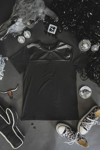 A LONESTAR SLATE *MIDNIGHT t-shirt from Western Edge Boutique, featuring a luxurious leather-like upper section, is laid flat on a dark surface. Surrounding the shirt are scattered disco balls, a pair of black and white sneakers, and shiny black decorations, suggesting a festive or party theme.